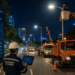 A professional cinematic shot of a modern street in Medan at night with bright LED street lights and professional maintenance crews pimpinan cherry picker trucks and digital circuit maps pimpinan bright and safe atmosphere pimpinan photorealistic pimpinan 8k resolution.