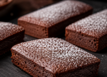 Foto deretan kue chocolate financier elegan berbentuk persegi panjang dengan taburan gula halus di atas papan batu tulis gelap,
