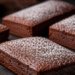 Foto deretan kue chocolate financier elegan berbentuk persegi panjang dengan taburan gula halus di atas papan batu tulis gelap,