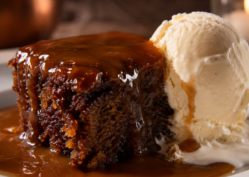 Foto sajian sticky toffee pudding hangat yang disiram saus karamel mengkilap beserta es krim vanila di atas piring keramik putih,