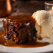 Foto sajian sticky toffee pudding hangat yang disiram saus karamel mengkilap beserta es krim vanila di atas piring keramik putih,