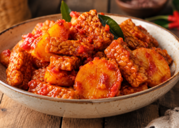 Foto sajian tempe dan kentang bumbu balado pedas mengkilap di dalam mangkuk keramik di atas meja kayu tradisional,