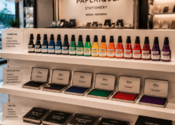 Jajaran botol tinta stempel premium berbagai warna dan alat bak stempel logam minimalis yang tersusun sangat rapi di dalam etalase toko perlengkapan stasioneri kantor
